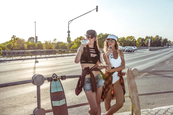 Dua gadis pacar siswa minum air di kota musim panas, papan seluncur, celana pendek baju renang. Beristirahat panjang rambut kecokelatan sosok. Background jalan mobil. Gaya busana, gaya hidup modern. Ruang kosong untuk teks . — Stok Foto