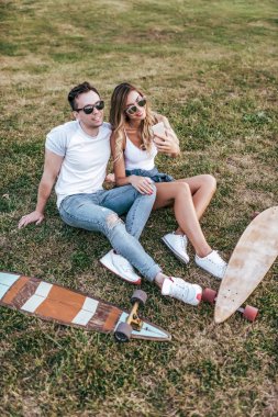 Genç çift adam kadın çim yaz şehir hafta sonu, mutlu olanlar eğlenmek şaka gülümseyen oturmak. Kaykay tahtası Longboard, gündelik giyim, aşk ilişkisi kavramı. Tarih toplantısı sevgi dolu arkadaşlar.
