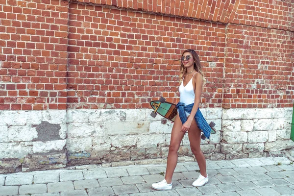 Happy girl walks summer city, smiling rests weekend, in hands skateboard board. Free space for text. Emotions joy fun, background wall. Tanned fitness figure athlete on longboard. — Stock Photo, Image