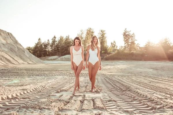 Duas meninas namoradas caminham na areia branca, em uma pedreira, junto ao mar, na praia do lago. Estilo de vida da moda, juventude moderna. Uma figura bronzeada. Estilo de vida ativo de uma mulher. Espaço livre . — Fotografia de Stock