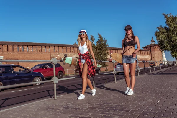 Två flickor flick vänner skateboard, sommar stad, promenader längs vägen, bakgrund av bilen, långt hår för Longboarding. Mode är livsstilen för ungdomar. Koncept idéer för en helg flykt bilen. Ledigt utrymme. — Stockfoto
