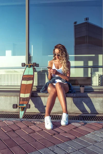 Gadis muda cantik kota musim panas, latar belakang bangunan kaca jendela, skateboard, jeans bodysuit putih, kacamata hitam. Menulis telepon, online aplikasi online jejaring sosial, membaca . — Stok Foto