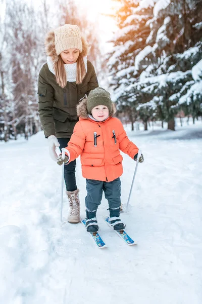 年轻的母亲保险支持孩子滑雪板, 3岁的小男孩儿子。冬季森林公园, 背景雪飘树。概念照顾孩子, 第一步体育和健身训练. — 图库照片