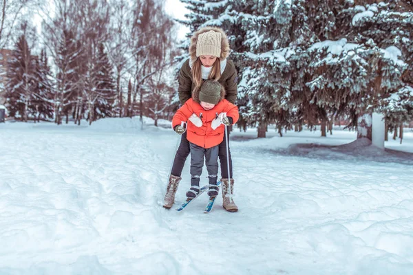 Junge Mutter versichert Kinderski, kleiner Junge Sohn 4 Jahre. Winter Waldpark, Hintergrund Schneeverwehungen Bäume. Konzept Betreuung Kind, erste Schritte Sport und Fitnesstraining. — Stockfoto
