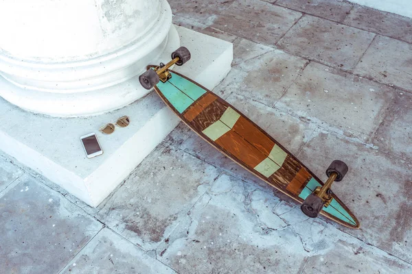 Longboard monopatín, se encuentra en la ciudad en verano en la columna blanca, el fondo es una baldosa vieja. Gafas de sol en oro derecha, teléfono móvil, smartphone. Estilo de vida activo, tendencias de estilo de moda . —  Fotos de Stock