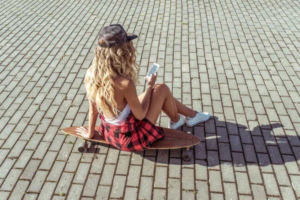 Blondes Mädchen mit langen Haaren, Sommerstadt sitzt Skateboard, liest Bewerbungen im Internet, soziale Netzwerke. Mode Lifestyle, moderne Ideen Konzeptentrends. braun gebrannte Figur. Hintergrund Pflastersteinstraße. — Stockfoto