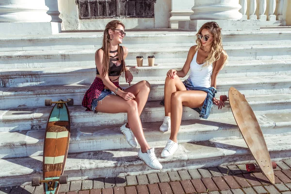 Dos chicas amigas, sentadas en las escaleras en verano en la ciudad, charlando, riendo feliz y sonriendo, monopatín, tazas de café, hermosas y bronceadas figuras. Concepto mejor amiga novia . — Foto de Stock