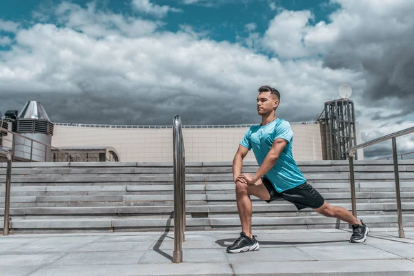 男性アスリートは、ジョギング、フィットネストレーニング、スポーツウェア、集中力と目標に焦点を当てる前に筋肉を伸ばし、夏の街を肺にします。階段の背景青い空。テキストの空き領域. — ストック写真