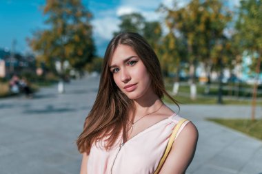 Elbiseli bir kızın yakın çekim portresi, şehirde yaz, uzun saçlı, esmer, doğaya yaslayan, arka plan ağaçları, mavi gökyüzü, ilginç bir görünüm, omuzçanta