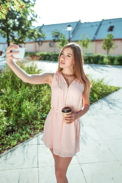 Chica en vestido, verano en la ciudad, se para en el teléfono en la mano, foto selfie en el teléfono inteligente, aplicación, redes sociales en línea, videollamada por Internet, fotografías a sí misma charlando taza de café de té — Foto de Stock