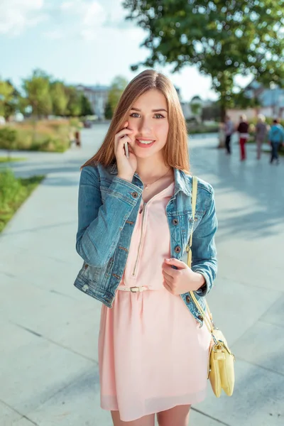 Beautiful student girl jeans, summer city, stands her hand phone, rings smartphone, smiles happy, emotions of pleasure relaxing and good phone conversation, pink dress, yellow bag over her shoulder