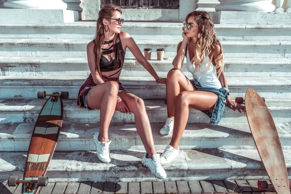 Δύο όμορφα κορίτσια φιλενάδες το καλοκαίρι στην πόλη, κάθονται στα σκαλιά. Σανίδες για πατίνια, φλιτζάνια καφέ. Μακριά μαλλιά μαυρισμένη σιλουέτα, γυαλιά ηλίου. Ιδέα συνάντηση καλύτερες αδελφές φιλενάδες. — Φωτογραφία Αρχείου