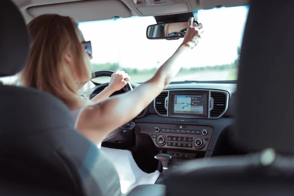 Ragazza che guida un'auto, si guarda allo specchietto retrovisore, corregge. Parcheggiare l'auto e guidare verso il retro, monitorare i passeggeri e i bambini in auto. Configura i comandi in auto . — Foto Stock