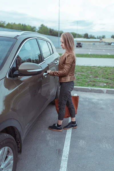 Menina de casaco e jeans abre a porta do carro no estacionamento perto de um shopping center. Activação do alarme. SUV crossover verde no verão na cidade. Retorno da loja com saco e compras . — Fotografia de Stock