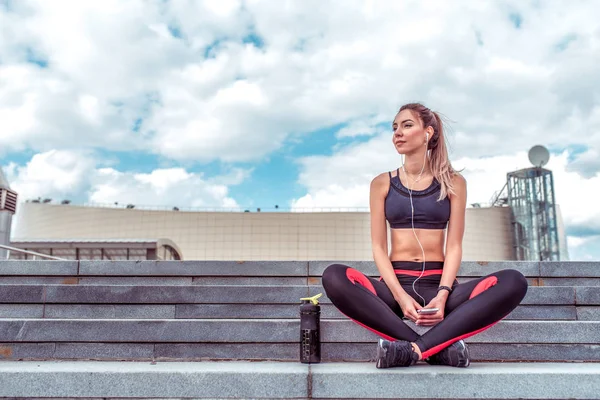 Güzel kız atlet, şehirde yaz aylarında bir egzersiz sonra oturur, rahatlatır, sokakta fitness, bir akıllı telefon müzik dinler, yaz aylarında doğanın tadını çıkarın, su ve protein ile bir shaker.