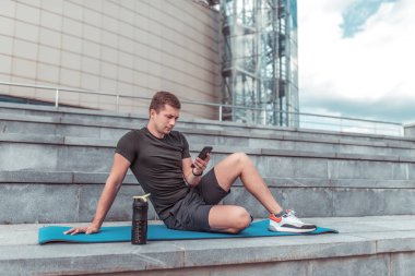 Bir telefon ile bir adam, şehirde bir eğitim oturumunda yaz aylarında, sokakta fitness, bir akıllı telefon, telefonda bir online uygulama, su ve protein ile bir shaker bir mesaj yazıyor.