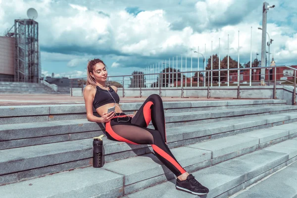 Una mujer disfruta relajarse, en sesión de entrenamiento de verano en la ciudad, escucha música con auriculares, con los ojos cerrados, fitness al aire libre, disfrutar de la naturaleza en verano, agitador con agua y proteínas . — Foto de Stock