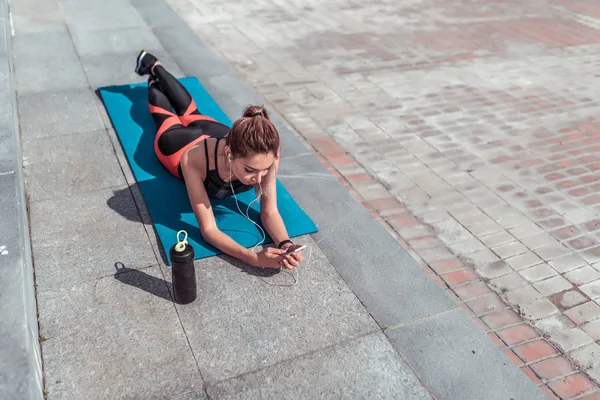 Спортсменка-дівчина, лежить на килимку для йоги після тренування, відпочиває в місті на фітнес, активний спосіб життя, шейкер з енергетичним напоєм та білком, онлайн-додаток для навушників у смартфоні, вільне місце . — стокове фото