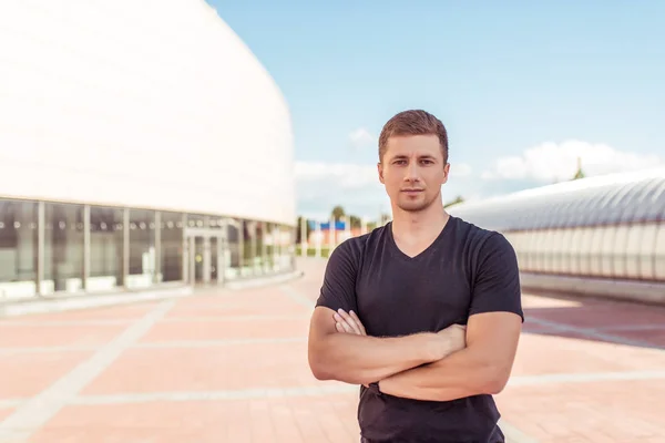 Atleta masculino, se encuentra en el verano en la ciudad contra el fondo del edificio, luce un estilo de vida activo, posando, camiseta negra, brazos musculares. Motivación y confianza —  Fotos de Stock