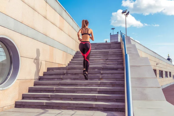 Kilátás a hátsó, nyáron a városban a lány fut fel a lépcsőn, az aktív életmód, az egészséges életmód fitness jellegű, sportruházat nadrágot felső, a háttérben a lépések, kék ég — Stock Fotó