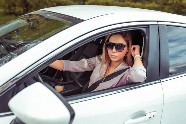 Mulher bonita dirigindo um carro, no verão na cidade, óculos de sol, terno rosa, figura bronzeada, close-up retrato, branco classe executiva sedan, estacionamento da cidade, mulher de negócios . — Fotografia de Stock