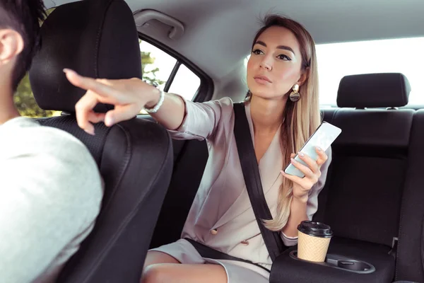 Taxisofőr mutatva ülés parkoló, üzleti nő azt mutatja, megáll a keze, ül az autóban, rendelkezik telefon, online app okostelefon, szigorú rózsaszín öltöny, hosszú haj, csésze kávé, üzleti taxi City nyár. — Stock Fotó