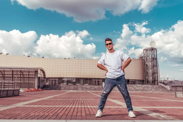 Jovem um dançarino em uma camiseta branca, jeans, posando para um líder, no verão na cidade, em óculos de sol, nuvens de construção de fundo, hip hop ativo, estilo de vida dos jovens, espaço livre para texto — Fotografia de Stock