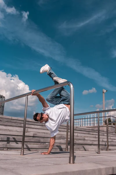 Young guy a dancer in a white T-shirt, jeans, dancing break dance in summer in the city, in sunglasses, white sneakers background building clouds, active hip hop, youth lifestyle — Stock Photo, Image