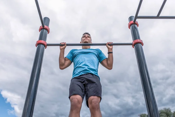 Mužský atlet se v létě ve městě vytahuje na vodorovný bar. Koncept síly, fitness cvičení ve městě. Sportovní oblečení, modré trička, mraky pozadí sportovní hřiště. — Stock fotografie