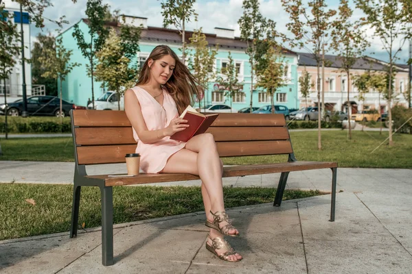 Una estudiante lee un libro de papel sentado en un banco en el verano en un parque de la ciudad. Feliz sonrisa, leyendo un libro de arte, una taza de café de té, espacio libre para el texto . — Foto de Stock