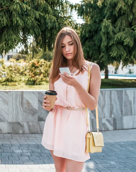Siswa perempuan dengan gaun merah muda, membaca pesan di telepon, aplikasi online di Internet. Secangkir kopi atau teh, kantong di atas bahumu. Musim panas di kota, pohon berlatar belakang cemara . — Stok Foto