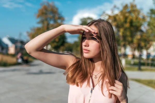 Siswa perempuan dengan gaun merah muda, menutup tangannya dari matahari yang cerah, melihat ke kejauhan, hari yang panas cerah. Di musim panas di kota . — Stok Foto