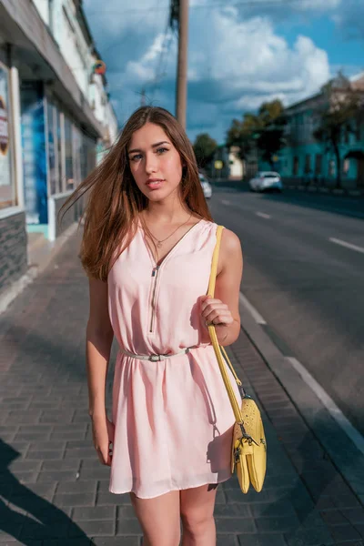 Belle fille en été dans la ville par la route. Cheveux longs, maquillage quotidien, robe rose, sac à bandoulière jaune. Nous attendons des amis sur le trottoir . — Photo