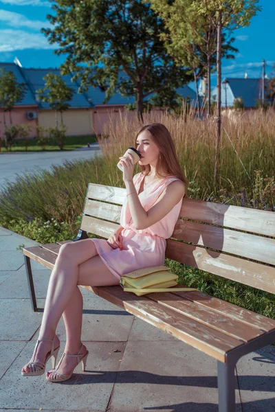 Seorang gadis duduk di bangku taman kota di musim panas, minum teh dari secangkir kopi, tas tangan. Istirahat setelah bekerja, makan siang saat makan siang, istirahat di tempat kerja, semak-semak pohon latar belakang, bangku kayu. Baju sandal bertumit pink . — Stok Foto