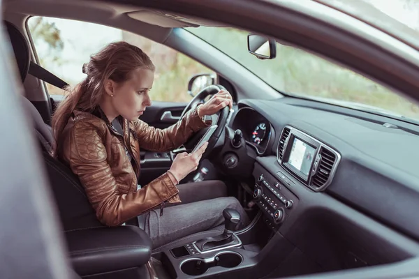 woman driving car, selects navigation on phone s display screen, turns voice assistant, searches for route to Internet, checks map with phone navigation in car. Leather jacket summer autumn on trip.