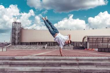 spor adam, bir kolunda duruyor, genç bir adam bir dansçı, bir alt mola, adım ve bir bina, kot pantolon, güneş gözlüğü, beyaz bir t-shirt arka plan karşı şehirde yaz aylarında hareket dans