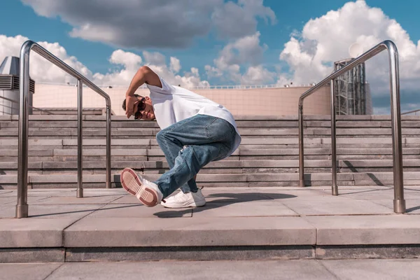 Ein junger Kerl ein Tänzer, macht eine untere Pause, tanzt im Sommer in Bewegung in der Stadt vor dem Hintergrund von Stufen und einem Gebäude, Jeans, Sonnenbrille, weißes T-Shirt — Stockfoto