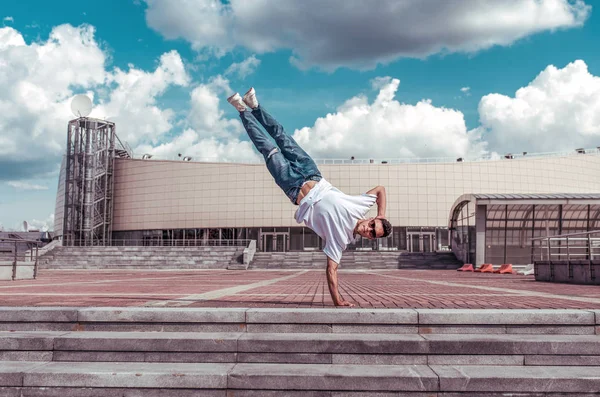 spor adam, bir kolunda duruyor, genç bir adam bir dansçı, bir alt mola, adım ve bir bina, kot pantolon, güneş gözlüğü, beyaz bir t-shirt arka plan karşı şehirde yaz aylarında hareket dans