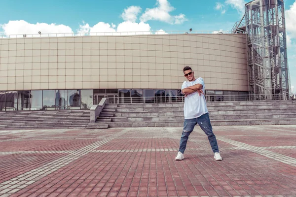 Sporty male dancer, standing in pose of confidence, young guy, free space for text of moticavia, in summer in city against the background of steps and a building, jeans sunglasses, white T-shirt — Stock Photo, Image