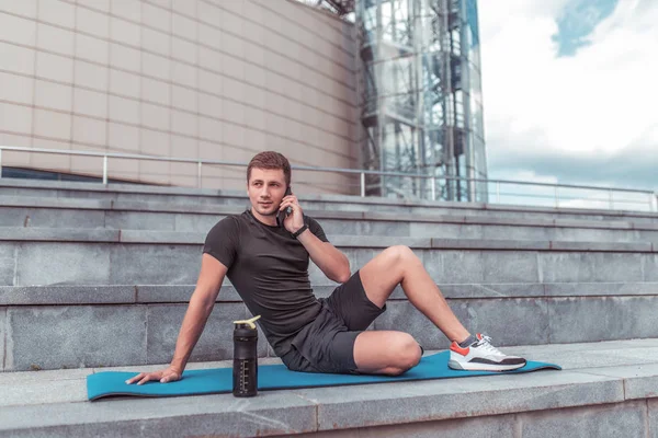 Erkek atlet, şehirde yaz, bir yoga ceket istirahat, telefonla arama, spor şort ve t-shirt, su ve protein ile shaker, bir akıllı telefon online arama uygulaması giyiyor.