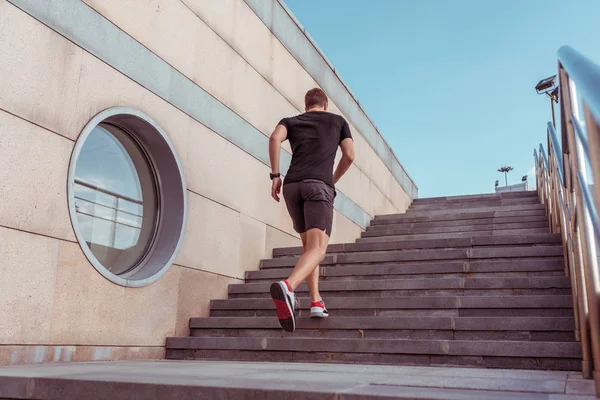 後ろから見える男性アスリートは、夏の市内の階段を走り回る。スポーツウェアTシャツショートパンツ。フィットネス、若者のライフスタイル、市内の健康的なライフスタイル。テキストの空き領域. — ストック写真