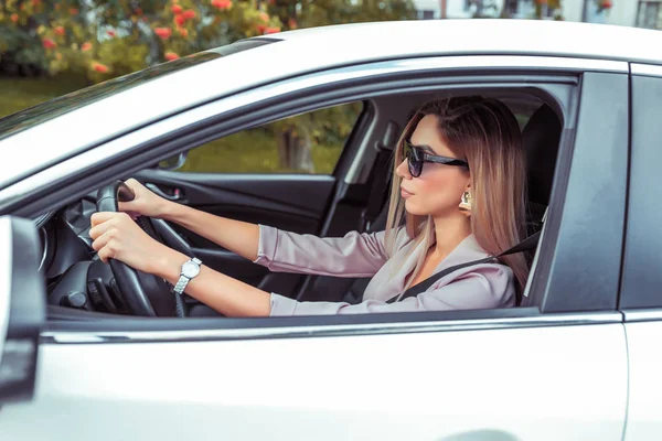 Mulher de negócios bonita senhora dirigindo um carro em óculos de sol, no verão na cidade, um terno formal bege, couro bronzeado, brincos de ouro, um relógio de marca caro em sua mão . — Fotografia de Stock