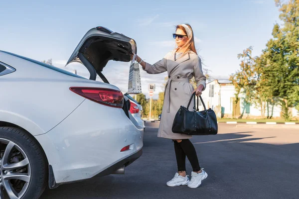 woman parking lot shopping center, opens trunk car, puts bags with purchases. Black bag with package. Concept shopping trip, parking in summer in city. In casual clothes in the fall.