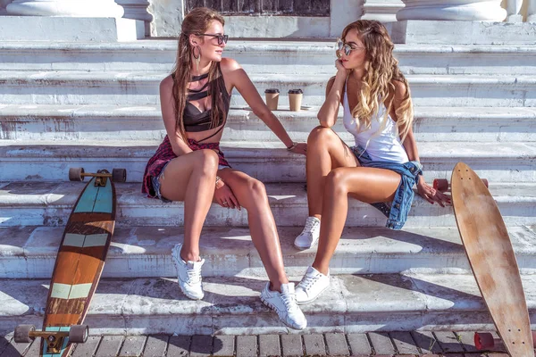 Schöne Freundinnen, die sich auf den Stufen des Instituts unterhalten, Tassen Kaffee mit Tee, Longboard-Skateboards, Kommunikation am Wochenende, Studentenleben, Sonnenbaden, Relaxen. — Stockfoto