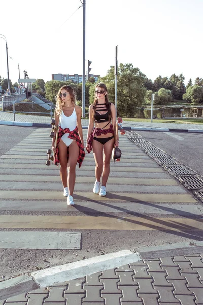 Two girlfriends walk in summer in city, background cross the road, swimsuits, shorts and baseball caps, skateboard long sunglasses. Weekend walk, youth lifestyle, modern style.