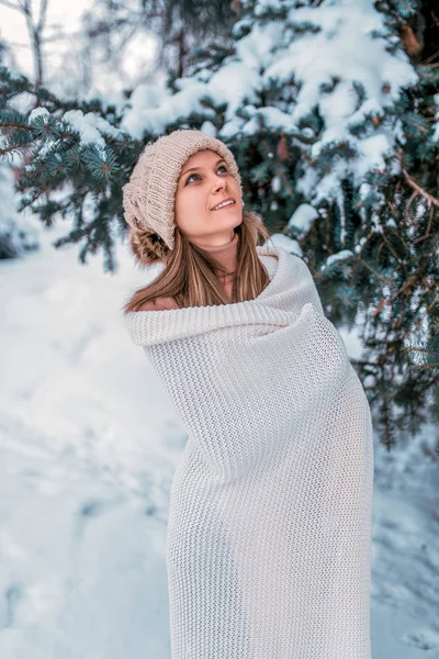 Donna felice inverno con un cappello bianco. Avvolto in una coperta calda, sfondo, neve, cumuli di neve, albero di Natale verde. Emozioni della gioia di una pausa weekend in vacanza . — Foto Stock