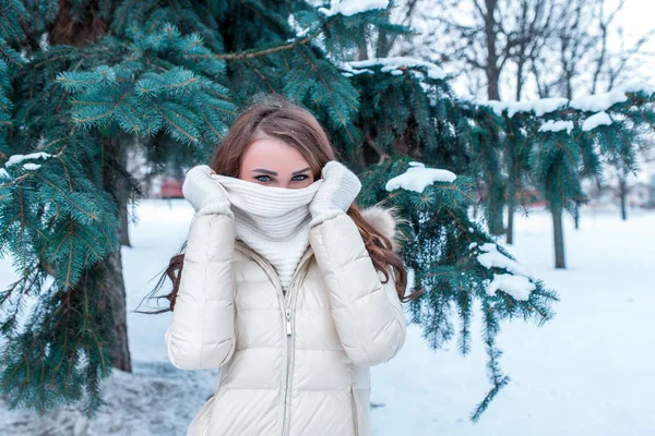 Dívka v zimě zakrývá její tvář šátkem, v zimě šťastně vyschlé na pozadí zasněženého zeleného smrku. Odpočívej v zimních prázdninách v letovisku. Bílé palcové a teplé sako. — Stock fotografie