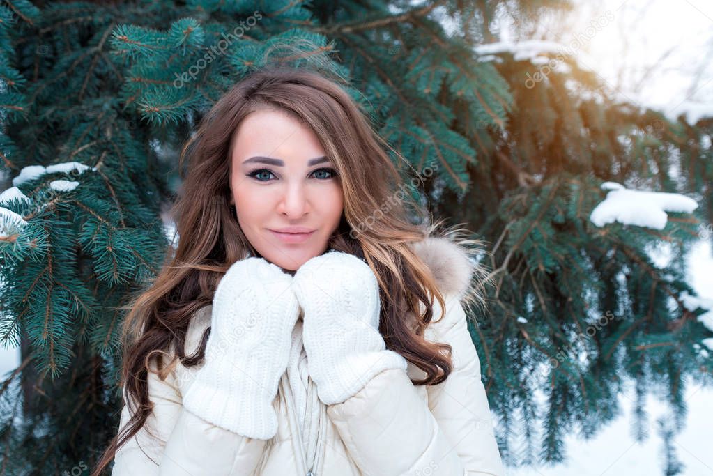 Portrait of a girl in winter on background of green branches of spruce, happy warms herself in white warm mittens. Emotions of comfort of a weekend break in a winter resort.
