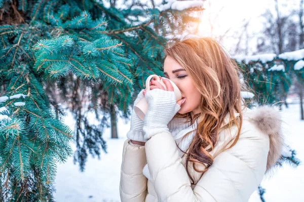 woman in winter outdoors in park, drinking coffee, tea from mug, beautiful long hair, dying winter snowy green spruce. Rest in winter holidays at resort. White mittens and warm jacket.