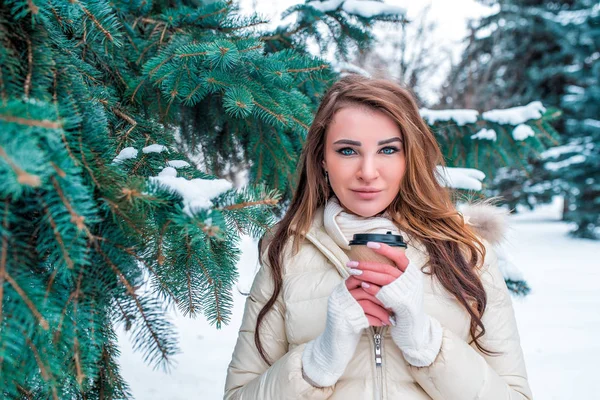 Mulher no inverno no parque de rua, aquece-se com xícara de café, chá, cabelo longo bonito, seca inverno abeto verde nevado. Descanse em férias de inverno no resort. mitenes brancas e casaco quente . — Fotografia de Stock